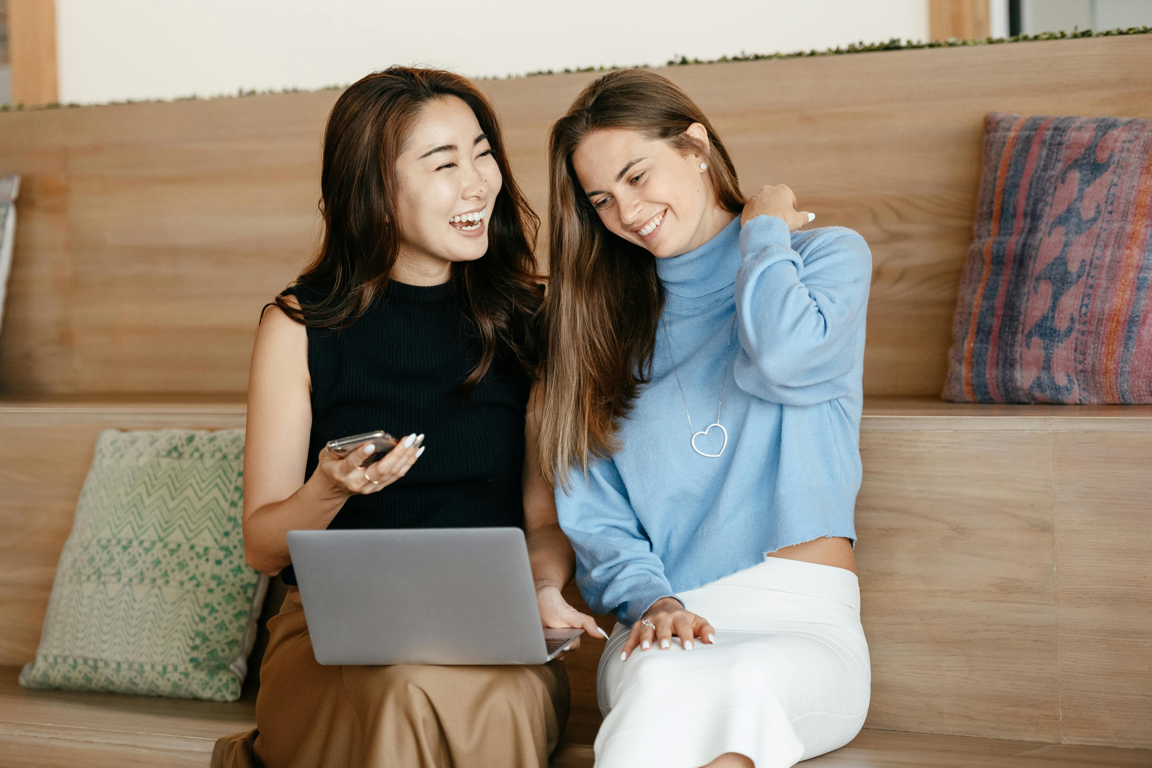 Two woman sitting next to each other, holding a computer while laughing.