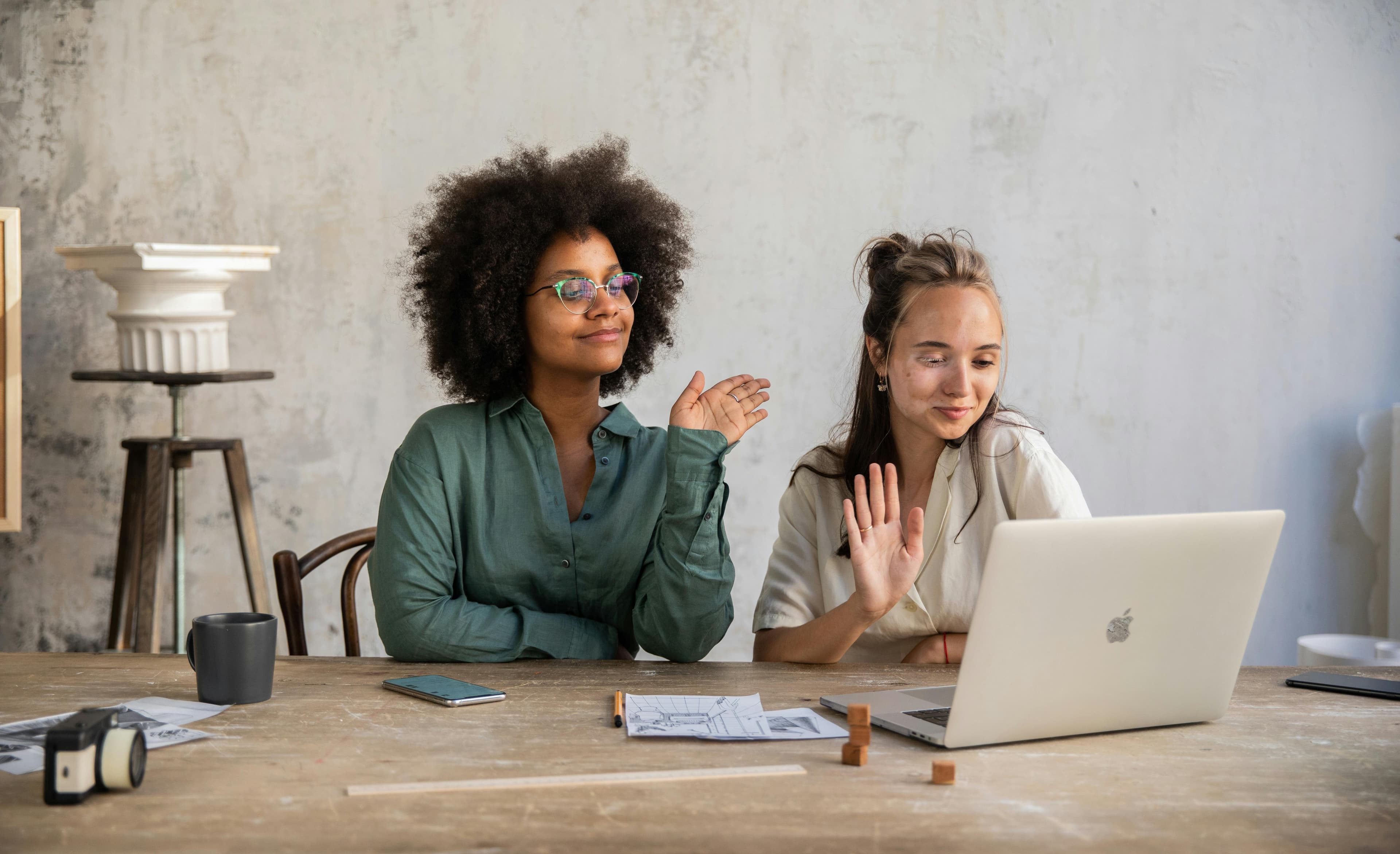 Two women sitting at a desk, smiling and waving at a laptop.